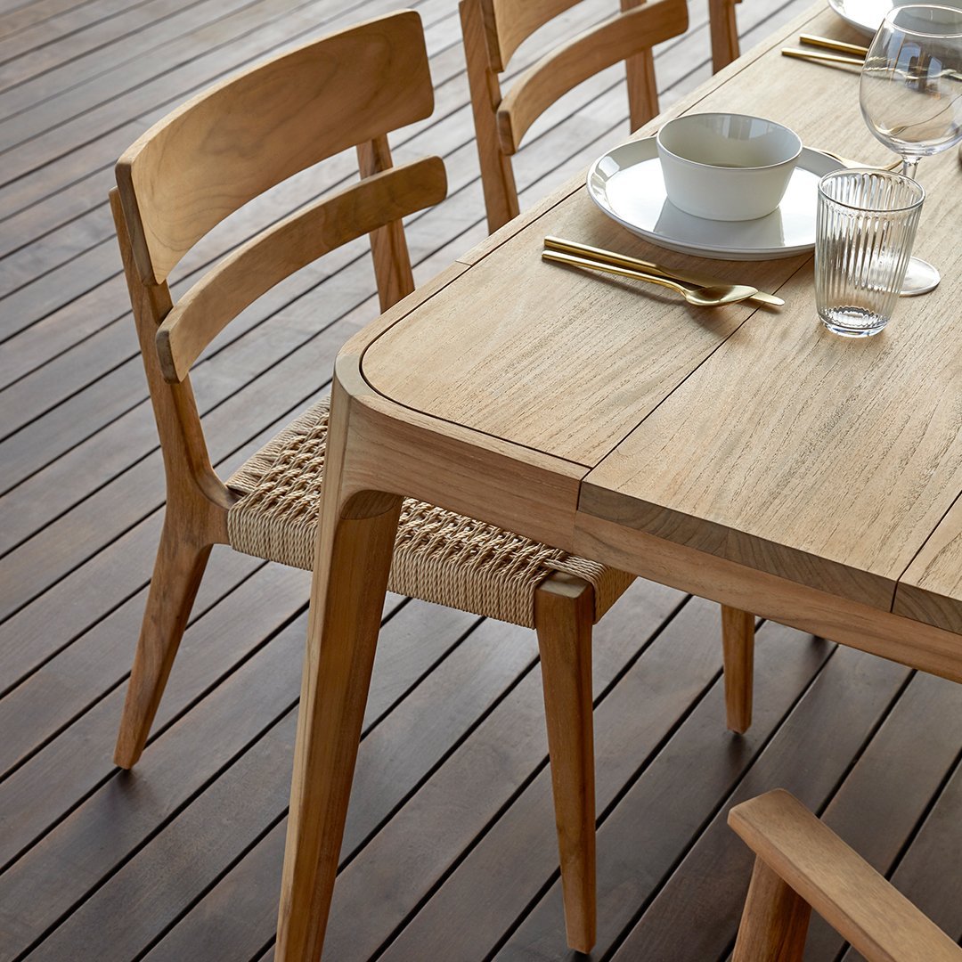 Stylish tan dining table with woven seats, set against a serene outdoor backdrop of greenery and clear skies.