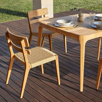 Light wood dining table with natural wicker-patterned chair, set against a green lawn and wooden deck.