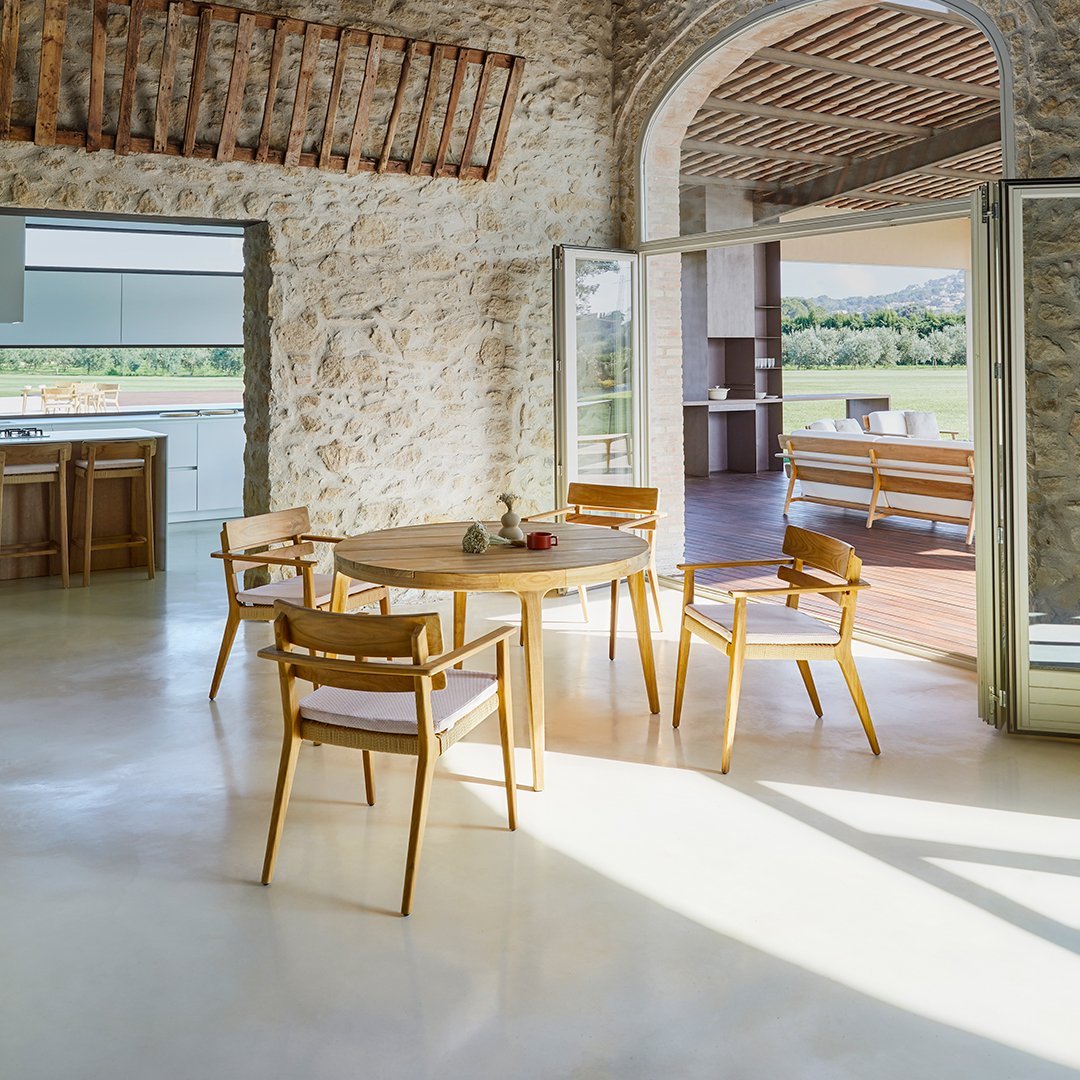 Wooden dining table with light-toned chairs, natural stone walls, and warm, earthy color palette in a bright indoor space.
