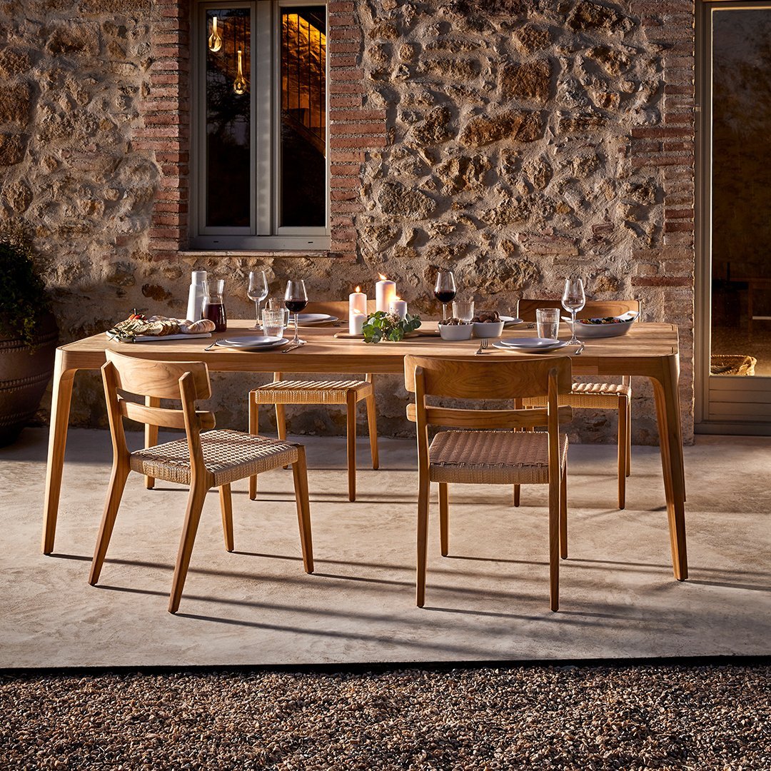 "Natural wood dining table and chairs with a warm finish, set against a textured stone wall. Elegant outdoor decor."