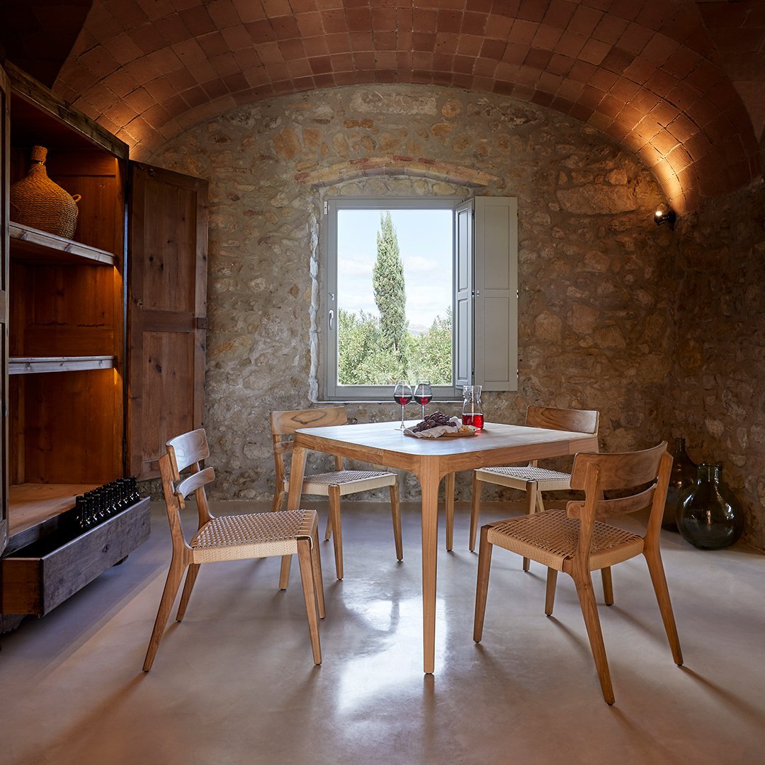 Light wooden dining table and chairs with neutral tones, set in a rustic stone-walled room with natural light.
