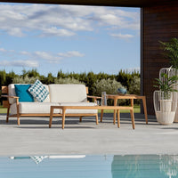 "Beige outdoor sofa with blue geometric cushion, paired with a light wood table and green potted plant against a blue sky."