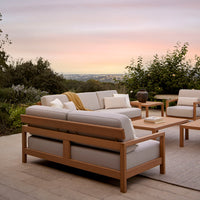 Light gray cushions with a warm wood frame, complemented by a neutral outdoor rug against a sunset backdrop.