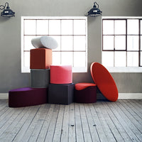 Colorful stacked cushions in various shapes, featuring shades of red, pink, gray, and purple against a light wood floor.