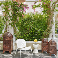 "White chair, rust-colored lanterns, vibrant green foliage, and pink blooms create a colorful outdoor setting."