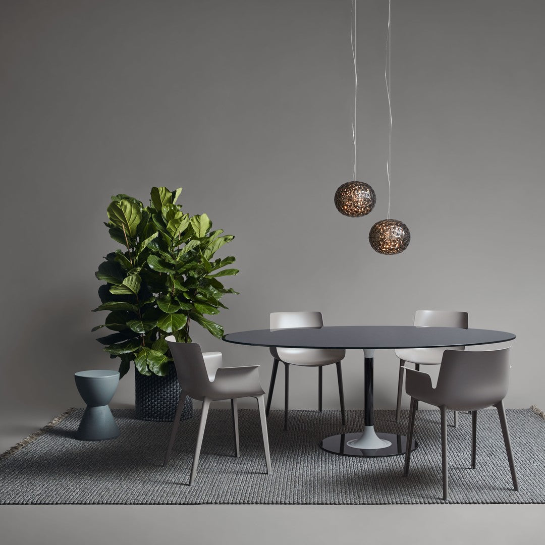 Modern dining setup with gray chairs, a round dark table, black and gold pendant lights, and a vibrant green plant.