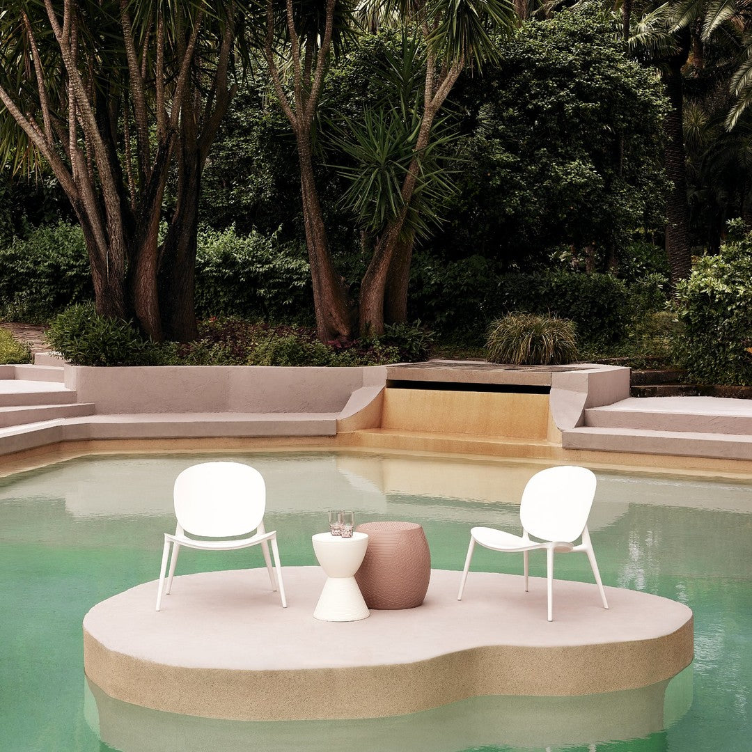 White chairs and a round, textured pink table on a light beige platform by a tranquil green pool amidst lush greenery.