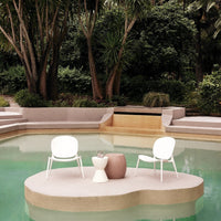 White chairs and a round, textured pink table on a light beige platform by a tranquil green pool amidst lush greenery.
