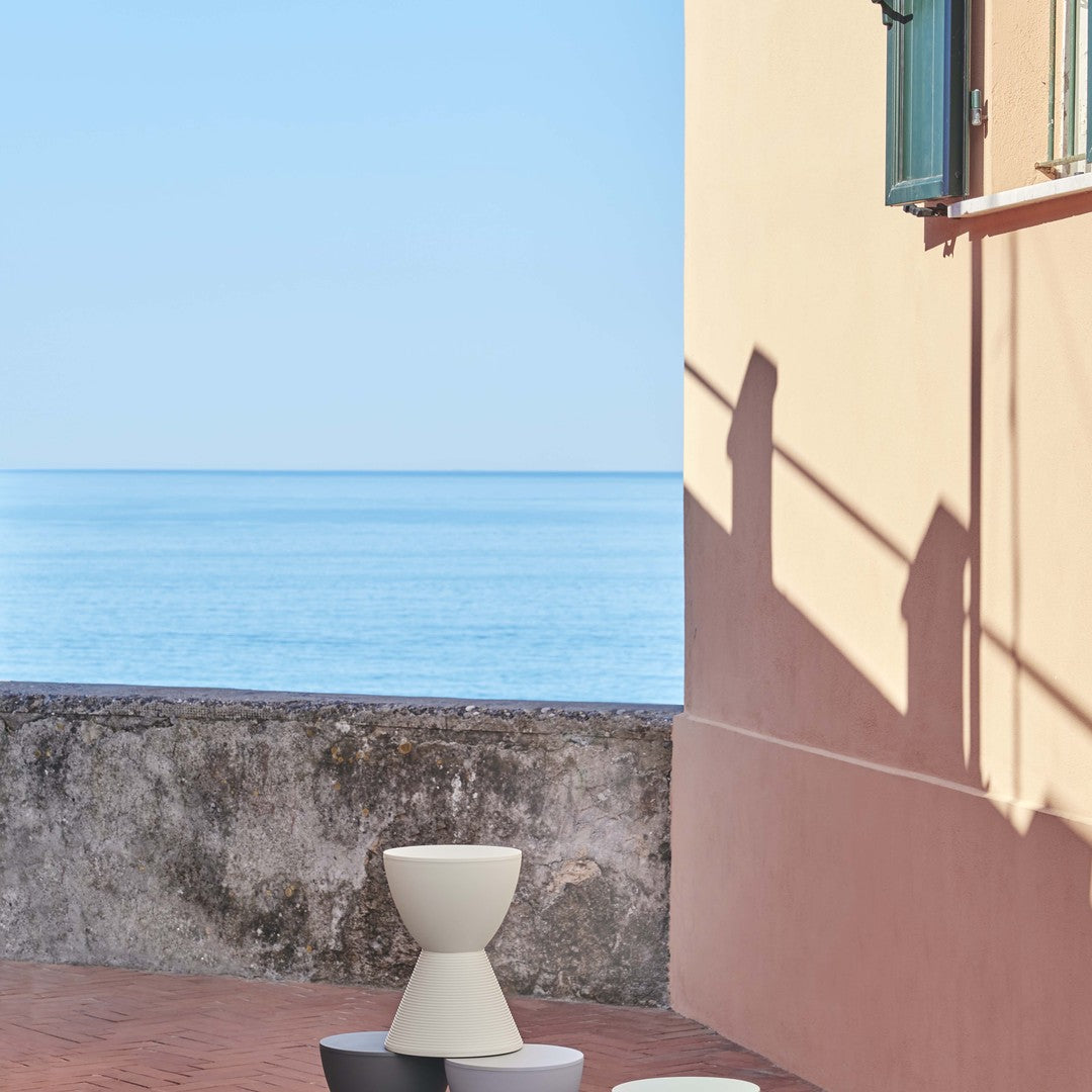 Three modern stools in white, gray, and black against a pastel pink background by a tranquil blue sea.