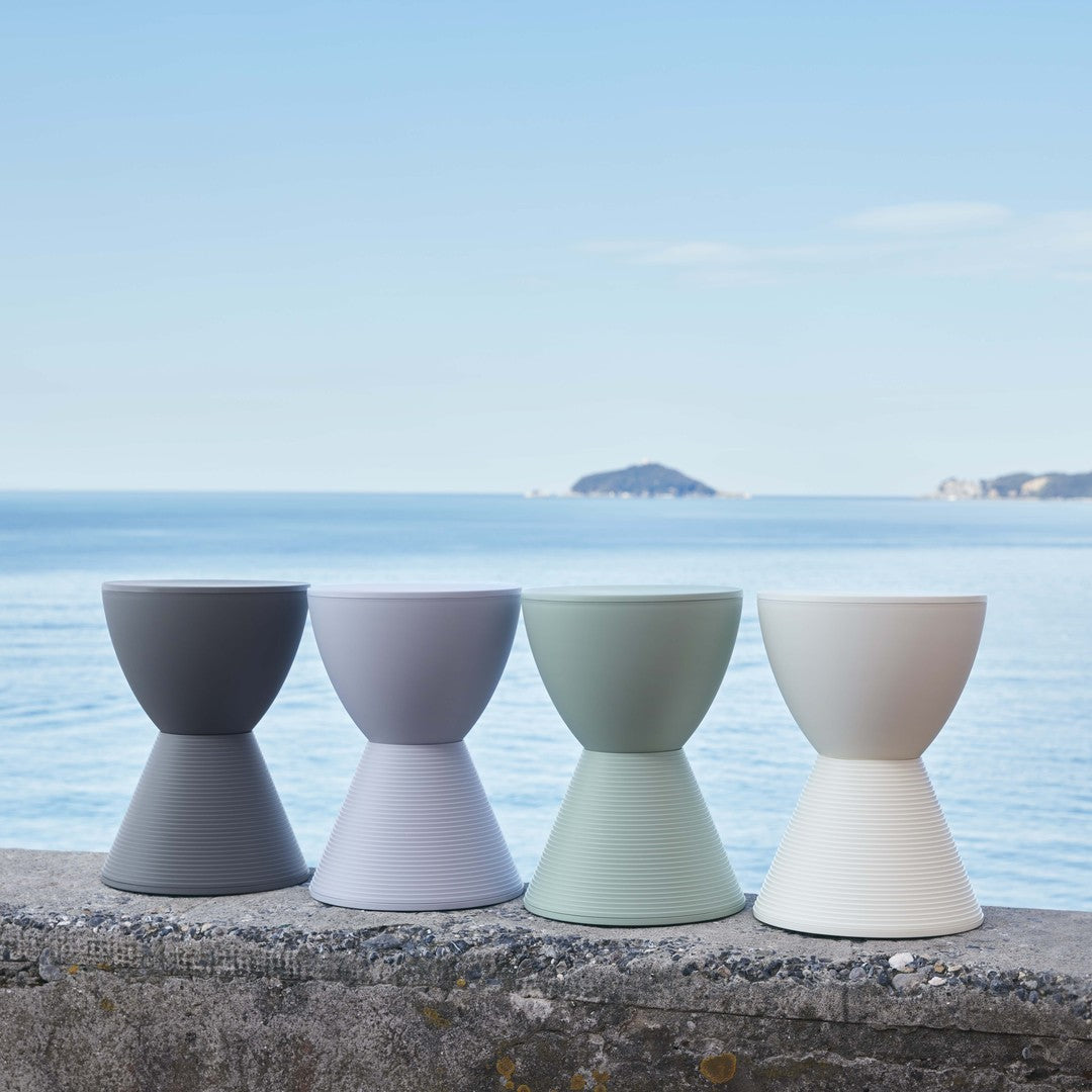 Four stylish hourglass-shaped planters in charcoal, light gray, mint green, and ivory against a serene seascape.