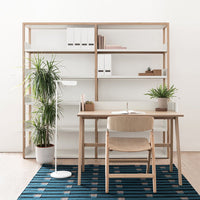 "Modern office setup with a white and wood bookshelf, blue striped rug, and light wood desk and chair."