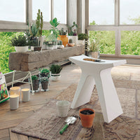 Sleek white stool on a rustic floor with earthy, patterned decor and lush green plants in bright natural light.