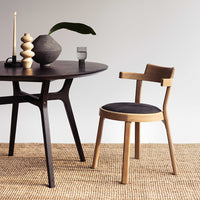 Natural wood chair with a black cushion, paired with a dark round table and neutral woven rug. Subtle, modern textures.