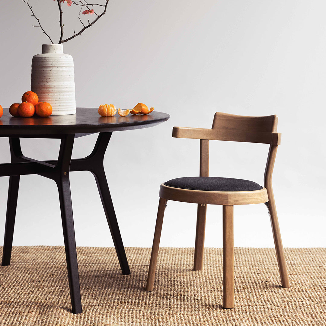 Modern wooden chair with a black upholstered seat, paired with a round dark table and neutral-toned woven rug.