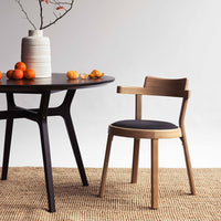 Modern wooden chair with a black upholstered seat, paired with a round dark table and neutral-toned woven rug.