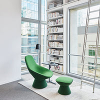 Green upholstered chair and matching stool against a bright, modern interior with large windows and bookshelves.