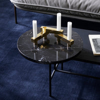 Round black marble coffee table with gold candle holder, accented by a dark blue carpet and light grey sofa.