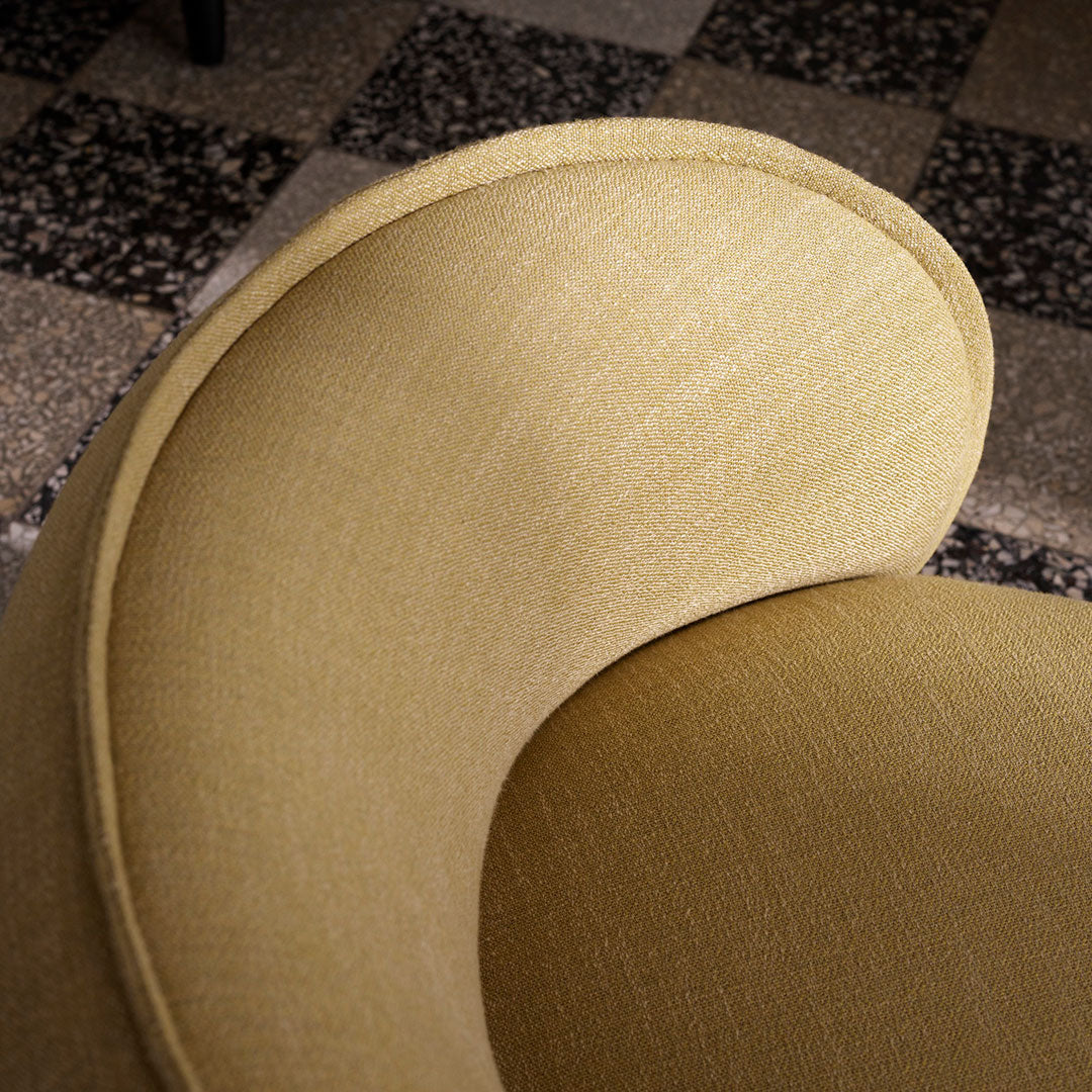 Curved chair in soft mustard yellow fabric with subtle texture, set against a patterned gray tiled floor.