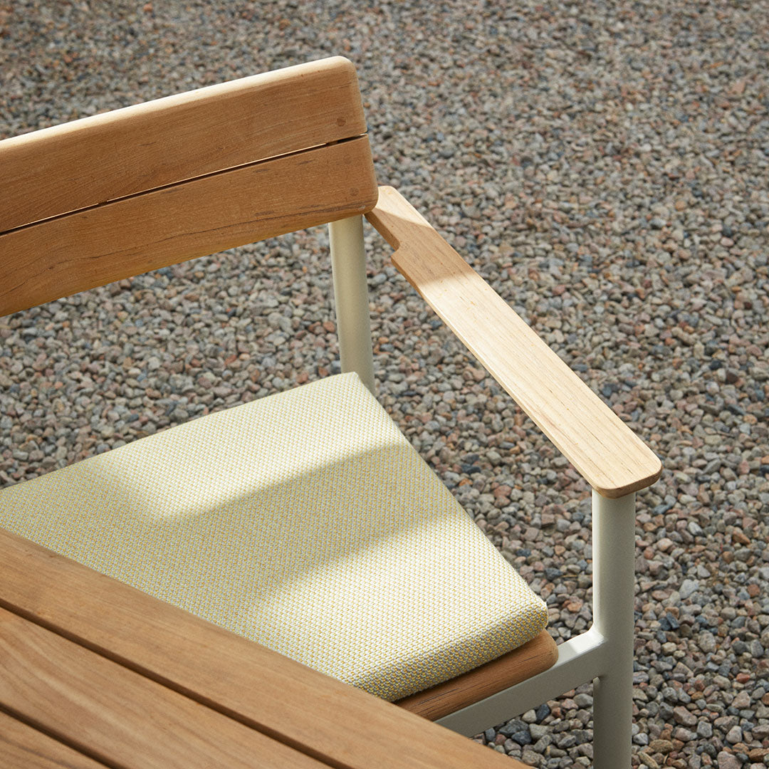 Light yellow cushion on a modern wooden chair against a textured gravel background. Minimalist design and earthy tones.