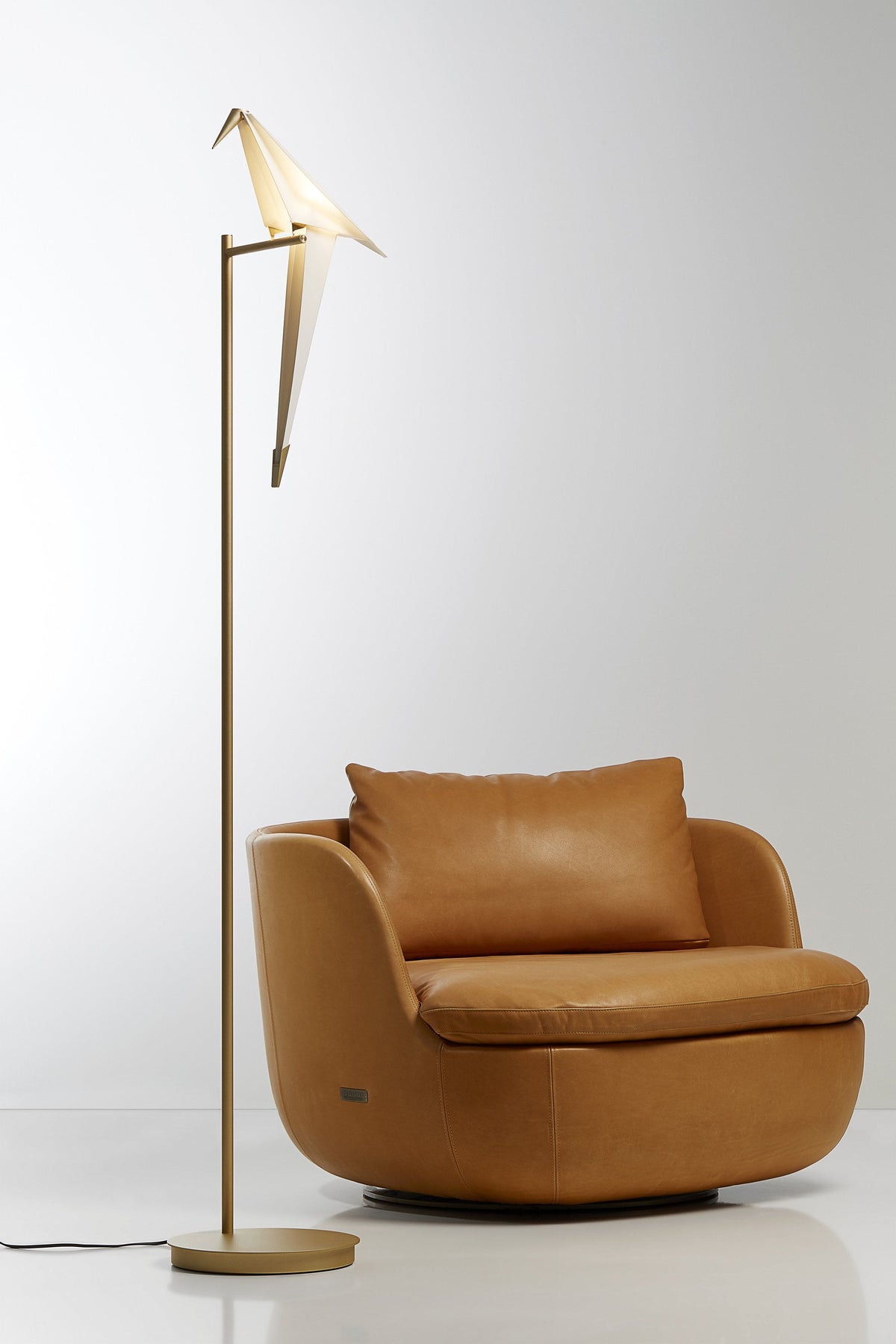 Modern gold floor lamp with a beige shade beside a smooth, tan leather armchair. Stylish and minimalist design.
