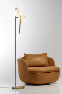 Modern gold floor lamp with a beige shade beside a smooth, tan leather armchair. Stylish and minimalist design.