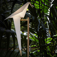 Modern white origami-style lamp with a sleek gold stand, surrounded by lush green tropical foliage.