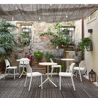 Outdoor patio scene featuring white chairs and round tables on a dark wooden deck, surrounded by lush greenery.