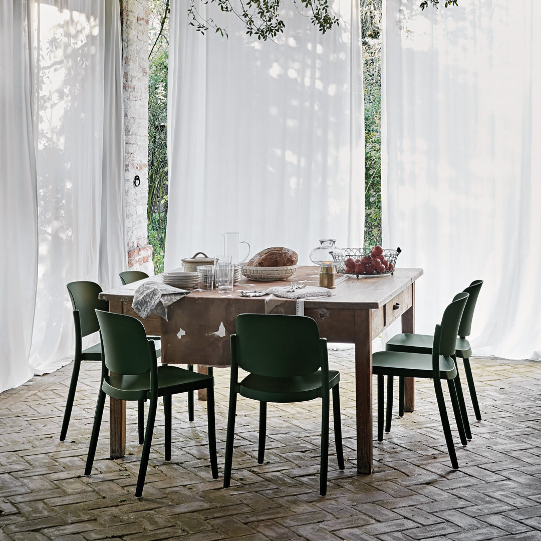 "Wooden dining table with rustic finish and green chairs, complemented by white drapes and natural surroundings."