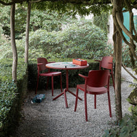 Red outdoor table and chairs in a green, leafy garden with a gravel surface, creating a vibrant yet natural setting.