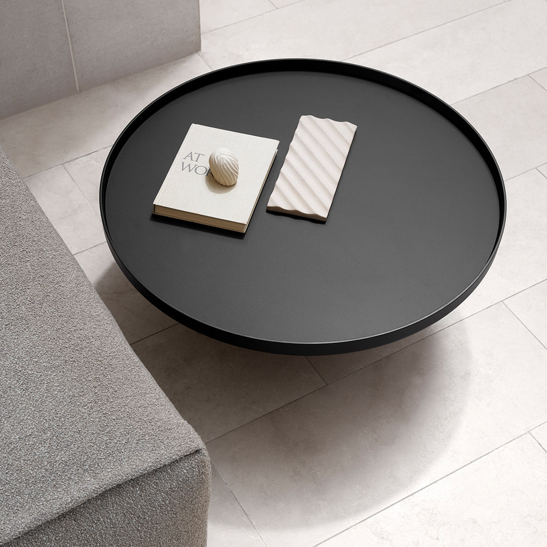 Round black coffee table with smooth surface, surrounded by light-colored tiles and a gray sofa. Simple, modern design.