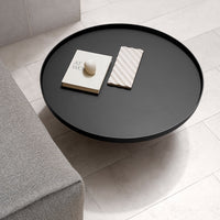 Round black coffee table with smooth surface, surrounded by light-colored tiles and a gray sofa. Simple, modern design.