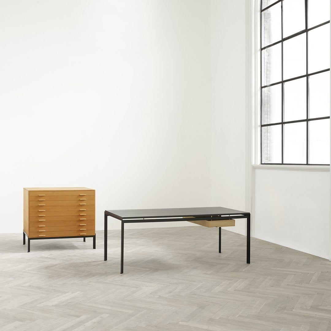 Modern black glass table with sleek black legs, paired with a warm honey-colored wood chest, set against a light gray herringbone floor.