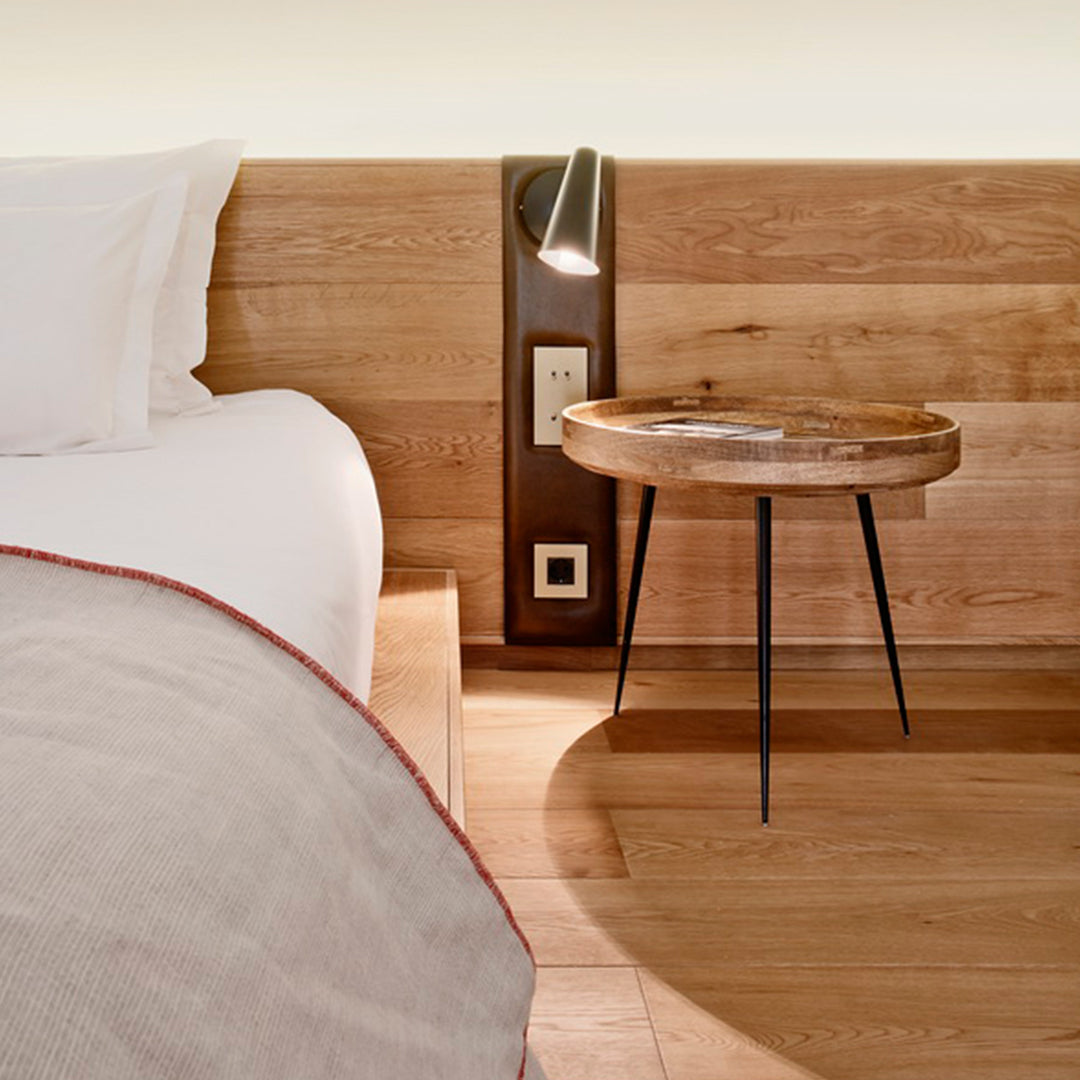 Wooden bedside table with black legs, soft cream bedding, and warm wood tones in a cozy modern bedroom setting.
