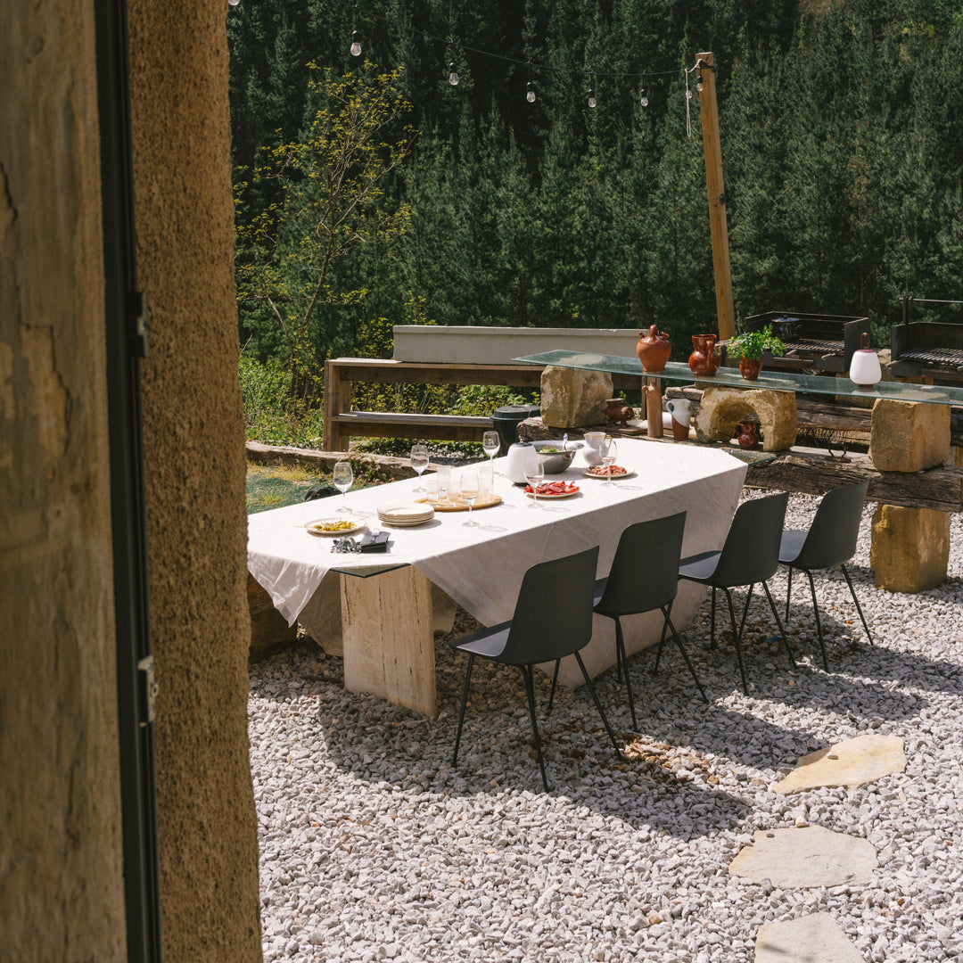 Stylish gray dining chairs around a rustic stone table, set against a backdrop of green trees and pebbled ground.