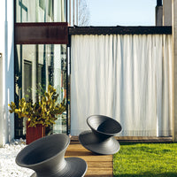 Black decorative chairs with smooth curves, contrasted by a white curtain and green foliage in a modern outdoor setting.