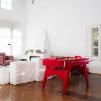 Red foosball table with colorful player figures, set against a bright, white interior with wooden flooring.
