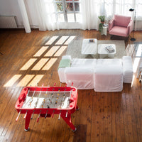 Red foosball table in a bright room with wooden floors; white couch, pink chair, and neutral area rug complete the look.