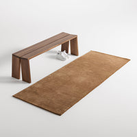 Rust-colored rectangular rug with a smooth texture, paired with a minimalist wooden bench on a neutral background.