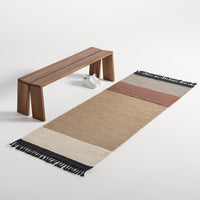 Striped beige, brown, and cream rug with black fringes, placed beside a sleek wooden bench.