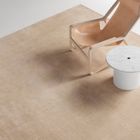 Beige rug with subtle texture, paired with a modern, transparent chair and a white marble side table.