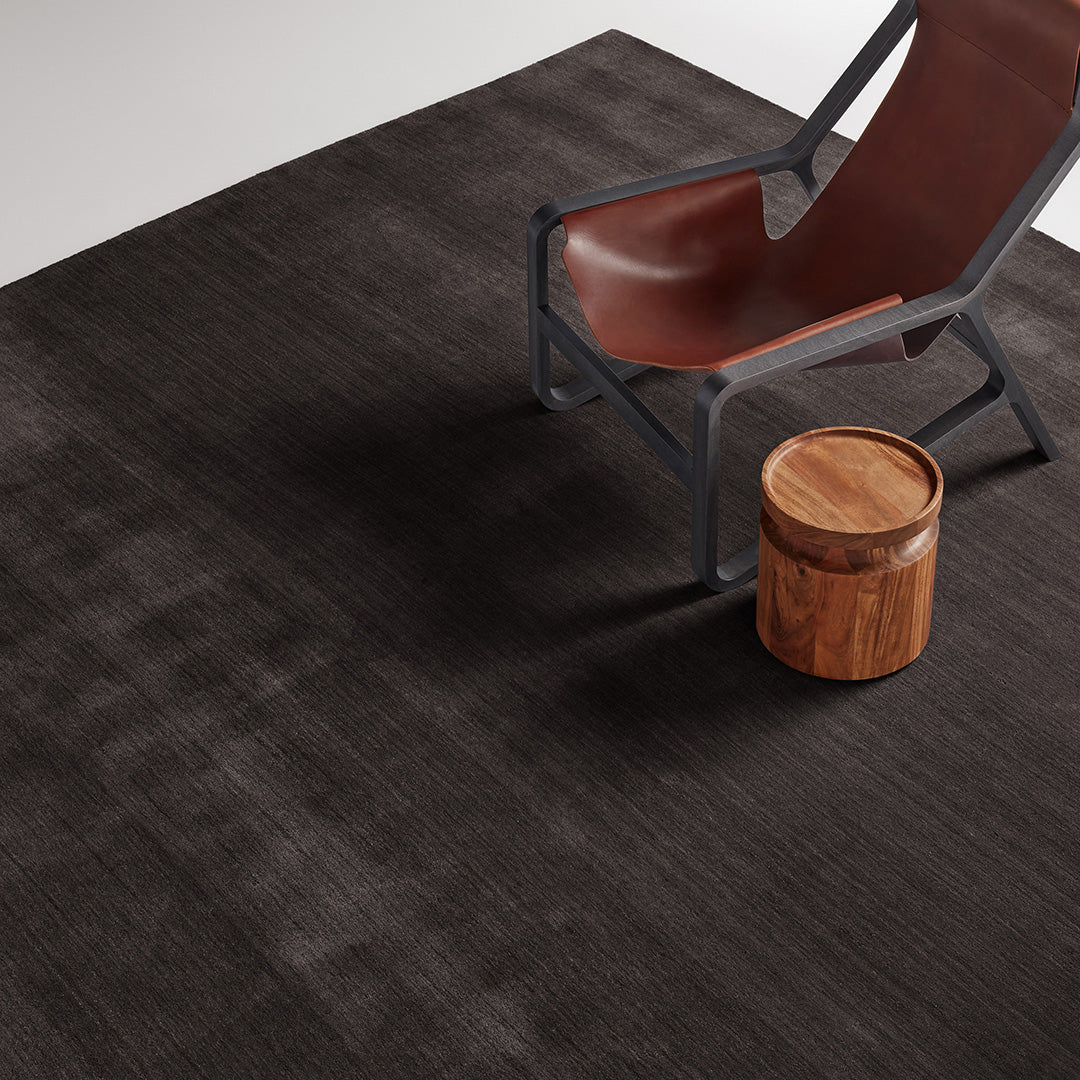Dark brown textured area rug with subtle pattern, paired with a modern chair and wooden stool.