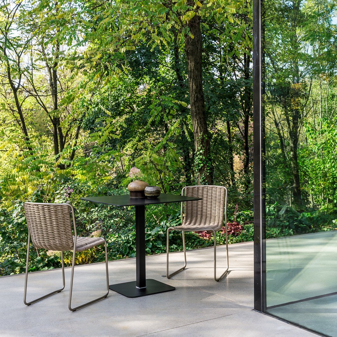Modern patio setup with neutral woven chairs, a sleek black table, and lush green surroundings in natural sunlight.