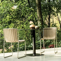 "Modern outdoor table set with beige woven chairs and a sleek black pedestal base, surrounded by lush greenery."
