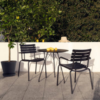 Modern black metal chairs with horizontal stripes beside a round table, set against lush greenery and a light-colored wall.