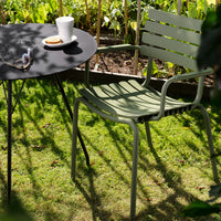 Olive green slatted chair beside a round black table, set against lush green grass in a garden.