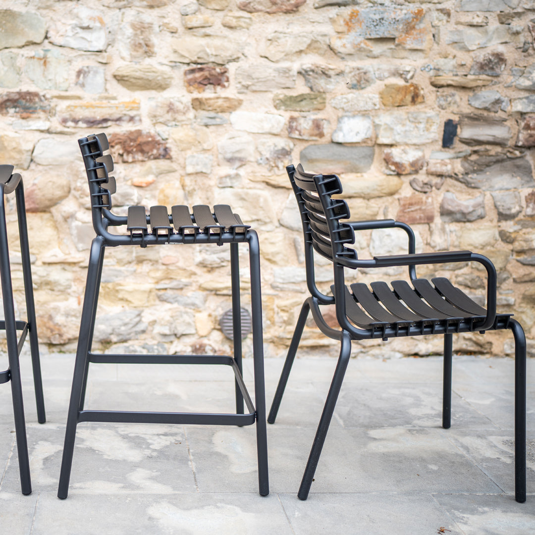 "Black metal chairs with slatted backs and seats against a rustic stone wall backdrop."