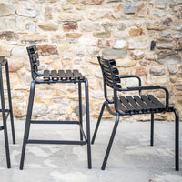 "Black metal chairs with slatted backs and seats against a rustic stone wall backdrop."