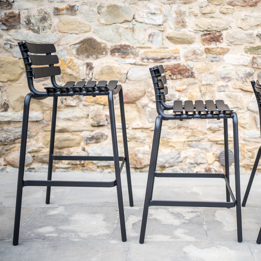 Black bar stools with slatted backs against a textured stone wall, showcasing a modern and minimalist design.
