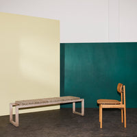Beige and dark green backdrop with a natural wood bench and chair featuring a woven seat in a minimalist setting.