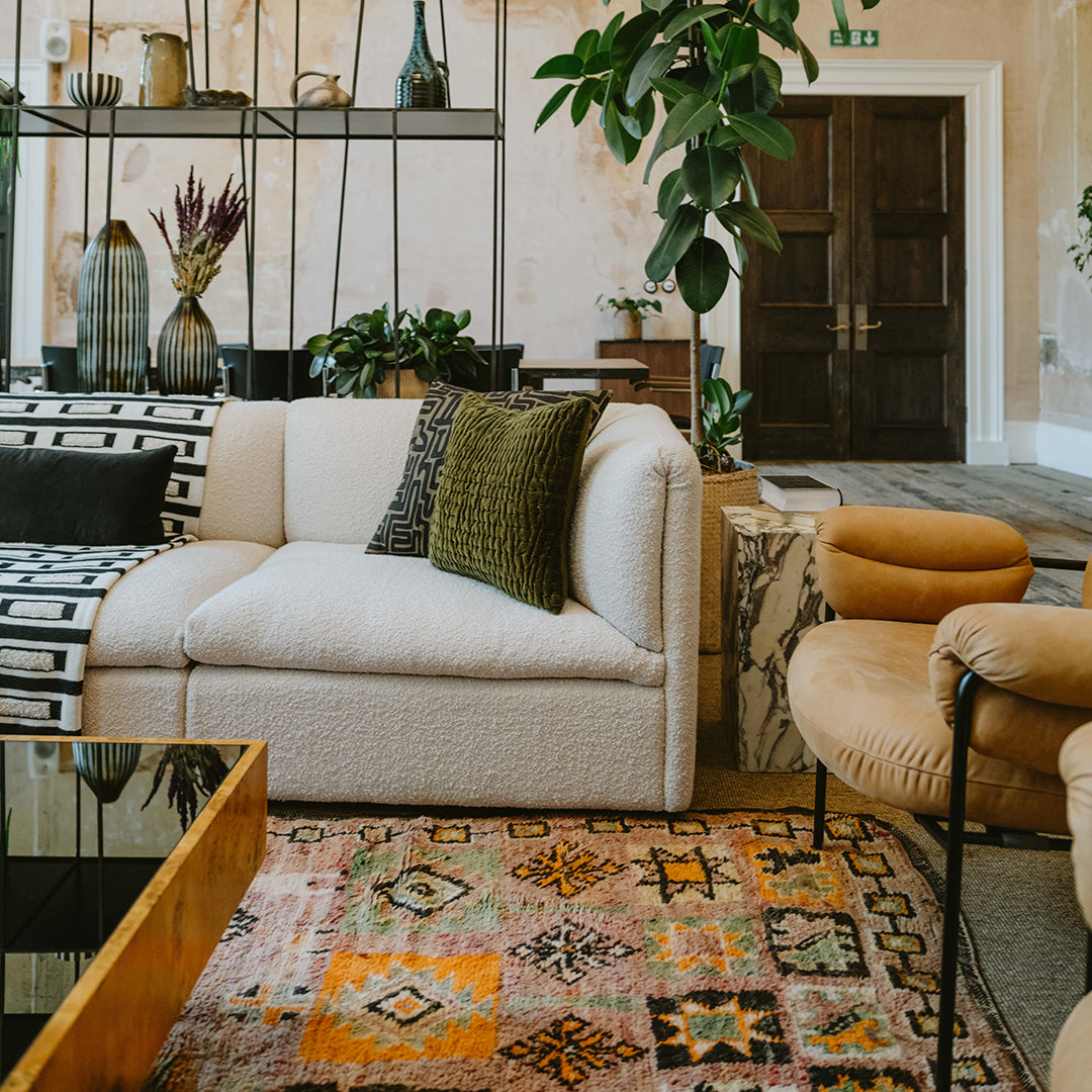 Neutral sofa with a dark green pillow, layered over a colorful, geometric-patterned rug in warm hues.